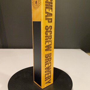 May include: A wooden beer tap handle with the words "CHEAP SCREW BREWERY" engraved vertically. The handle has a light wood finish with a black stripe and a circular logo. The logo says "Screw Brewery Co. Est. 2019".