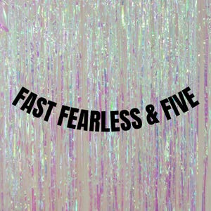 May include: A black banner with the text "FAST FEARLESS & FIVE" against a background of iridescent, shimmering, pink and white streamers.