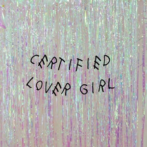 May include: A white background with iridescent, shimmering, plastic strips hanging from the top. The text "CERTIFIED COVER GIRL" is written in black across the middle of the image.