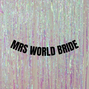 May include: A banner with the black text MRS WORLD BRIDE hanging in front of a shimmering, iridescent, light-reflecting fringe curtain. The vertical strips of the curtain create a metallic, multi-coloured effect with pink and blue tones.