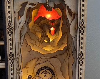 May include: A wooden diorama depicting a scene from The Lord of the Rings. A hobbit stands near a glowing doorway, while a fiery creature lurks in the shadows above. The diorama is lit from within, creating a warm and inviting atmosphere.