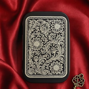 May include: A rectangular black metal tin featuring a detailed gold-colored engraved floral pattern with sunflowers and swirling vines on the lid, accompanied by a small, ornate antique-style brass key with a decorative bow and bit.