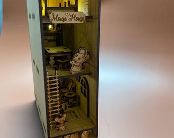 May include: A wooden mouse house with a sign that says "Mouse House" and a light inside. There are two mice figurines inside the house, one sitting in a chair and one standing on the floor. The house has a ladder, a table, and a chair. There are also some jars on the bottom shelf.