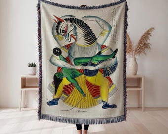 Könnte beinhalten: Eine gewebte Decke mit einer farbenfrohen Illustration einer mythischen Kreatur mit pferdeähnlichem Kopf, die eine grüne Figur hält. Das Kunstwerk hat eine Vintage-Ästhetik mit cremefarbenem Hintergrund und gefransten Kanten in Blau und Lila.