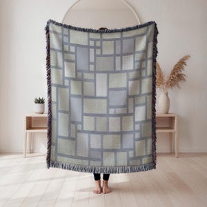 May include: A woven blanket with a geometric pattern of rectangles in shades of gray and green. The blanket has a fringed edge with hints of red and blue. The blanket is approximately 60 inches wide and 70 inches long.