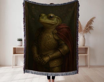 May include: A woven throw blanket featuring a regal frog in ornate golden armor and a red cape. The frog has large, expressive eyes and a serious expression. The background is a dark green with floral patterns, and the blanket has a fringed edge.