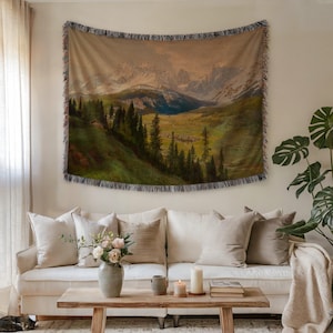 Puede incluir: Un tapiz tejido que representa un paisaje de montaña con un valle, árboles y picos nevados. El tapiz rectangular tiene un borde con flecos y está colgado en una pared sobre un sofá blanco con cojines beige. Una mesa de centro de madera y plantas están frente al sofá.