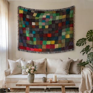 May include: A colorful, square-patterned throw blanket in shades of green, red, brown, and blue hangs on a wall. The blanket has a fringed edge and is displayed above a white sofa with neutral-toned pillows. A wooden coffee table and a potted plant are in front of the sofa.