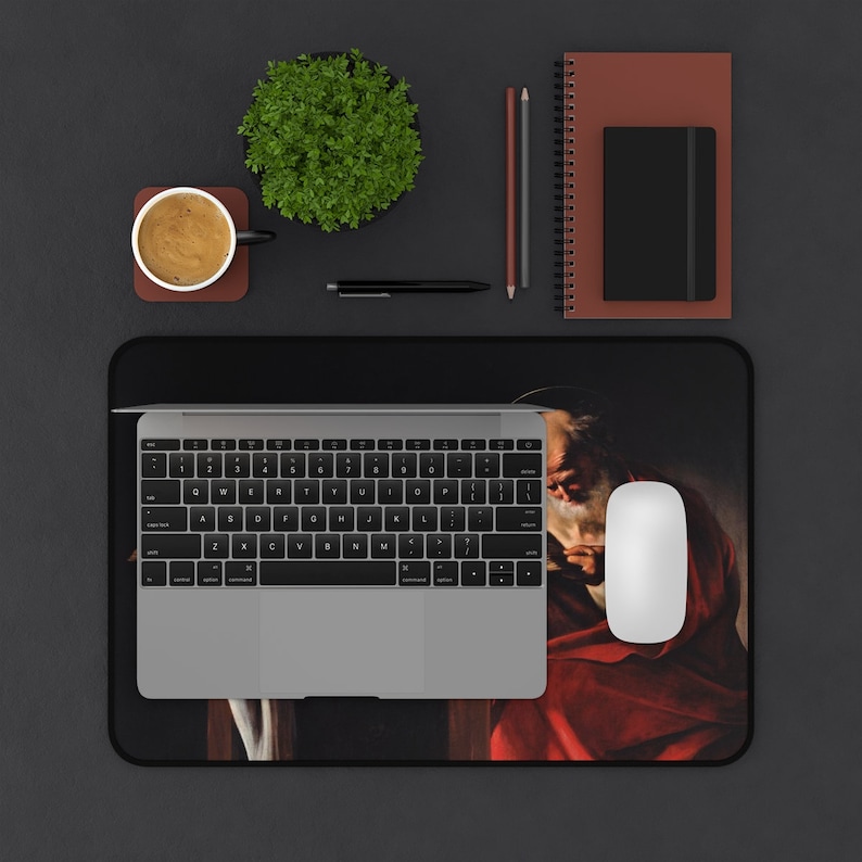 May include: A black mousepad with a painting of a man with a beard and a red robe. The man is holding a book in his hands. A laptop computer, a coffee cup, a plant, and a notebook are on a black surface.