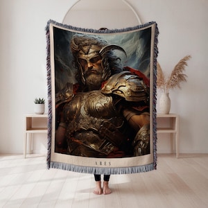 Könnte beinhalten: Eine gewebte Überwurfdecke mit einer detaillierten Illustration von Ares, dem griechischen Kriegsgott. Das Kunstwerk zeigt Ares in goldener Rüstung und Helm, mit dem Namen "ARES" unten. Die Decke hat einen Fransenrand und eine neutrale Farbpalette.
