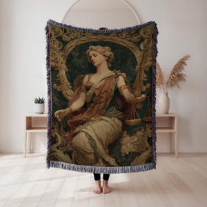 May include: A woven tapestry throw blanket featuring a classical painting of a woman holding scales. The artwork is framed by an ornate gold and green border. The throw has a fringed edge and is in shades of green, gold, and red.