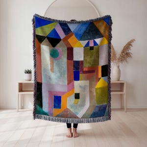 May include: A colorful woven throw blanket with a geometric design. The blanket features a variety of shapes and colors, including blue, green, yellow, and pink. The blanket has a fringed edge and is displayed in a room with light-colored walls and flooring.