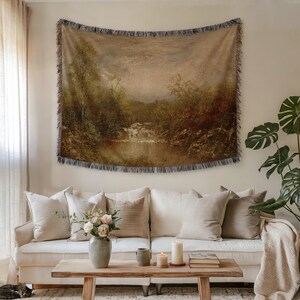May include: A woven tapestry featuring a landscape painting of a river and trees, displayed above a white sofa with beige pillows. The tapestry has a fringed edge. A wooden coffee table holds candles and a vase of flowers.
