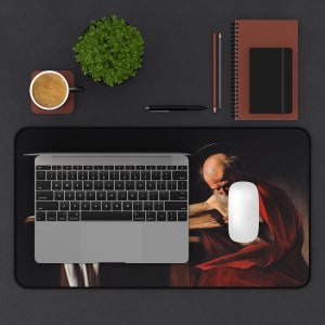 May include: A black and white mousepad with a painting of a man with a beard and a halo, wearing a red robe. The man is sitting at a table with a book in front of him.