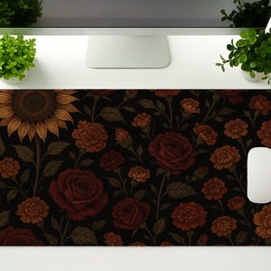 Puede incluir: Alfombrilla de ratón rectangular con un estampado floral de girasoles y rosas en tonos marrón, naranja y rojo sobre un fondo negro. Un ratón de ordenador blanco está a la derecha.