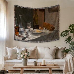 May include: A woven tapestry depicting a still life with bottles, a glass, and a loaf of bread. The artwork is in muted tones of brown, beige, and green. The tapestry is hanging above a white sofa with beige pillows, a wooden coffee table, and a vase of flowers.
