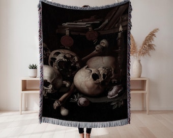 Puede incluir: Una manta tejida negra con una pintura de naturaleza muerta de calaveras, huesos y rosas. La obra de arte tiene una estética oscura y gótica con una vela y documentos antiguos. La manta tiene un borde con flecos.