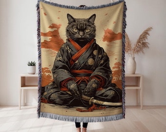 May include: A woven throw blanket featuring a black cat in a samurai outfit. The cat is depicted in a seated position, wearing a black kimono with red accents and holding a sword. The background has a beige and orange landscape design.