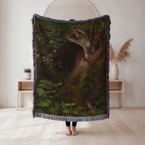May include: A woven throw blanket featuring a detailed forest scene with a large tree trunk and lush green foliage. The blanket has a dark color palette with a fringed edge, creating a cozy and artistic aesthetic.