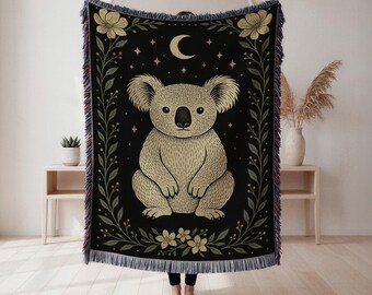 May include: A black woven throw blanket with a beige koala illustration, a crescent moon, and stars. The koala is surrounded by beige and green floral and leaf designs. The blanket has a fringed edge.