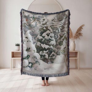 May include: A woven throw blanket featuring a winter landscape scene with snow-covered trees. The blanket has a fringed edge and a color palette of white, green, and blue. The blanket is held up by a person.