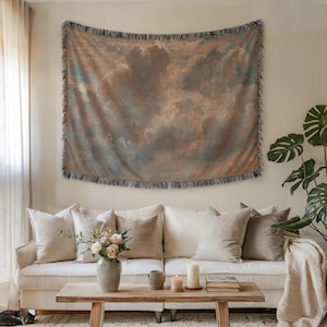 May include: A large tapestry with a cloudy sky design in shades of brown, blue, and white hangs above a white sofa. The tapestry has a fringed edge. The room is decorated with neutral-toned pillows, a wooden coffee table, and a potted plant.