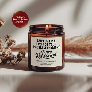 Puede incluir: Una vela de vidrio marrón encendida con una etiqueta blanca que dice "Smells Like It's Not Your Problem Anymore Happy Retirement". La vela es vegana y ecológica, hecha con una mezcla de cera de soja natural y una mecha de algodón. Un círculo rojo con texto blanco dice "Multiple Size & Scents Available."