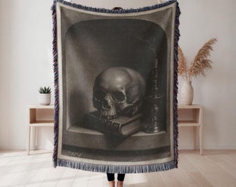 Puede incluir: Una manta tejida con una imagen en escala de grises de una calavera sobre un libro, una vela y un fondo oscuro. La manta tiene un borde con flecos con hilos azules, rojos y blancos. La obra de arte evoca un tema gótico o macabro.
