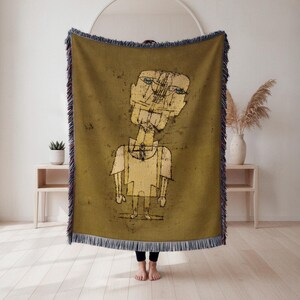 May include: Mustard-colored throw blanket featuring a stylized, abstract figure in yellow and black. The artwork has a distressed, vintage aesthetic. The blanket has a fringed edge and is held up by a person.