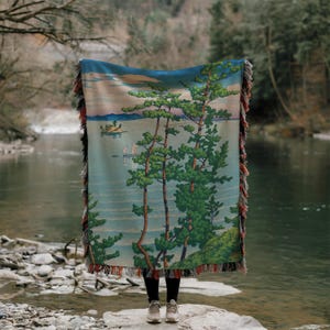 Könnte beinhalten: Eine gewebte Decke mit einer Landschaft mit Bäumen, Wasser und einem Boot. Die Decke hat eine Farbpalette aus Blau, Grün und Beige mit einem Fransenrand. Die Decke wird vor einer Person gehalten, die auf einem Felsen in der Nähe eines Flusses steht.