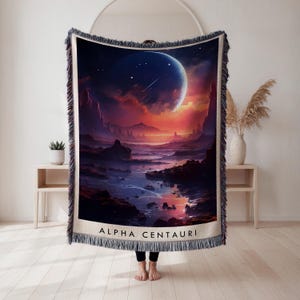 May include: Woven throw blanket featuring a vibrant space scene with a large planet, shooting stars, and a colorful sky. The design includes the text "ALPHA CENTAURI" at the bottom. The blanket has a fringed edge.