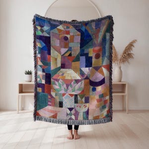 May include: A colorful woven throw blanket with a geometric abstract design. The blanket features a variety of shapes and colors, including blue, red, orange, and green. The blanket has a fringed edge and is displayed in a room with light-colored walls and wooden floors.