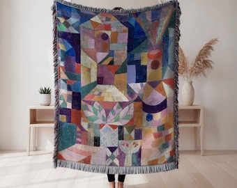 May include: A colorful woven throw blanket with a geometric abstract design. The blanket features a variety of shapes and colors, including blue, red, orange, and green. The blanket has a fringed edge and is displayed in a room with light-colored walls and wooden floors.