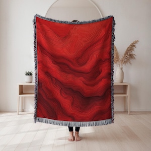 May include: A red throw blanket with a topographic map design. The blanket features a gradient of red hues, with darker shades at the bottom. It has a fringe border and is approximately 60 inches wide and 80 inches long.