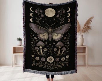 May include: Black throw blanket featuring a moth with outstretched wings, surrounded by floral and celestial elements. The design includes a full moon, crescent moons, flowers, and mushrooms. The blanket has a fringed edge.