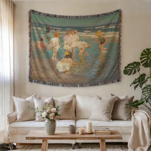 Puede incluir: Tapiz tejido que representa a niños jugando en la playa en un estilo vintage. La obra de arte presenta una paleta de colores apagados de azules, verdes y beige, con un borde con flecos. El tapiz se exhibe sobre un sofá color crema.