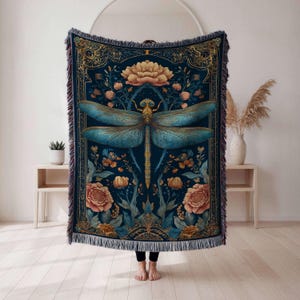 May include: A dark blue tapestry blanket featuring a large teal dragonfly with intricate floral and botanical designs. The blanket has a fringed edge and is decorated with gold accents and peach-colored flowers. The overall theme is nature and fantasy.