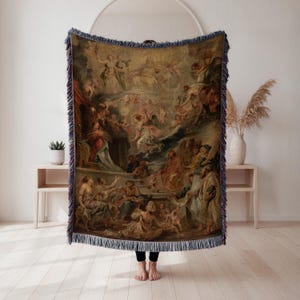 May include: A woven throw blanket featuring a classical painting with figures in shades of beige, brown, and gold. The artwork depicts a complex scene with many people and angels. The blanket has a dark blue fringe.