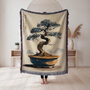 Könnte beinhalten: Eine beige Decke mit einer Illustration eines Bonsai-Baums in einem blauen Topf. Der Baum hat einen verdrehten Stamm und blau-grünes Laub. Die Decke hat einen Fransenrand und Text in der oberen linken Ecke.