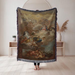May include: A woven throw blanket featuring a classical painting with figures in a cloudy sky. The artwork has a warm color palette with blues, yellows, and reds. The blanket has a fringed edge and is displayed indoors.