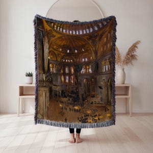 May include: A woven throw blanket featuring a detailed painting of a cathedral interior with a domed ceiling, arches, and figures. The blanket has a fringed edge with shades of brown, gold, and beige.