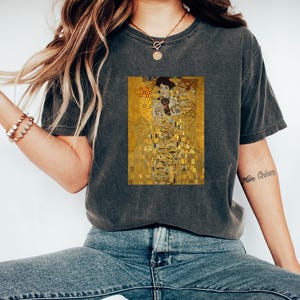 May include: A dark gray t-shirt with a print of a famous painting featuring a woman in a gold dress and a black cat. The painting is in a mosaic style with gold, yellow, and red colors.