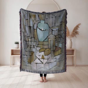 May include: A woven throw blanket featuring an abstract art design with a large blue vase as the central focus. The artwork is in shades of gray, blue, and yellow, with a fringed edge. The blanket is displayed indoors.