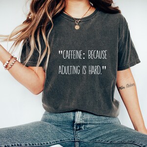 Puede incluir: Una camiseta gris oscuro con el texto "CAFFEINE: BECAUSE ADULTING IS HARD." impreso en blanco.