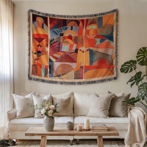 May include: A woven tapestry with a colorful abstract design of buildings and landscapes in shades of orange, red, and blue hangs above a white sofa. The tapestry has a fringed border. A wooden coffee table and decorative pillows complete the living room setting.