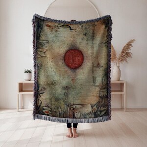 May include: A woven throw blanket with a unique, abstract design. The blanket features a large, red circular shape in the center, with a muted green and brown background. The edges are finished with a fringe.