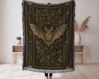 Puede incluir: Una manta verde oscuro con una ilustración detallada de un murciélago con las alas extendidas. El murciélago está centrado, rodeado de un diseño floral dorado. La manta tiene bordes con flecos.
