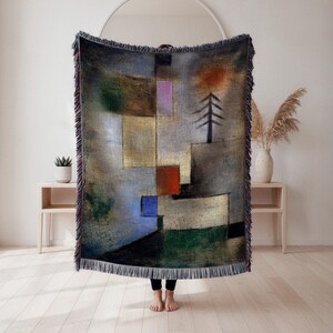 May include: A woven throw blanket featuring an abstract geometric design in muted tones of gray, beige, green, red, and blue. The artwork includes a stylized tree and a sun-like shape. The blanket has a fringed edge.
