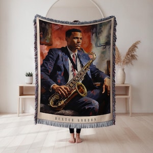 May include: Woven throw blanket featuring a portrait of Dexter Gordon playing a saxophone. The artwork depicts Gordon in a blue suit with a red tie, holding a gold saxophone. The bottom of the blanket has the name "DEXTER GORDON".