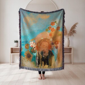 May include: A woven throw blanket with a colorful abstract design. The blanket features a central figure with a beard, surrounded by flowers in shades of orange, yellow, and white against a blue and gold background. The edges are fringed.